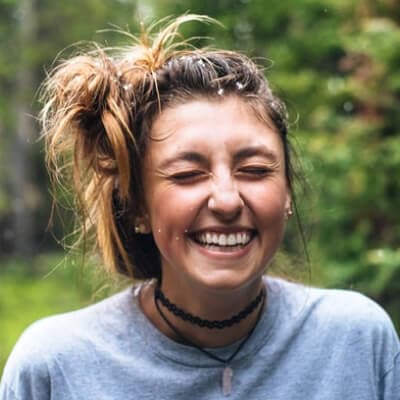 A young woman with a bright smile standing in a lush forest.